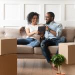 Couple sits on a beige couch amid unpacked boxes, smiling at a tablet together in a new home.