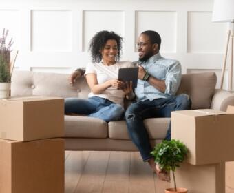 Couple sits on a beige couch amid unpacked boxes, smiling at a tablet together in a new home.