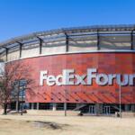 Exterior of the FedExForum arena with a red tiled facade and large silver ‘FedExForum’ letters against a bright blue sky.