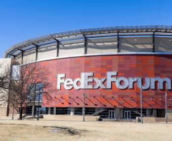 Exterior of the FedExForum arena with a red tiled facade and large silver ‘FedExForum’ letters against a bright blue sky.