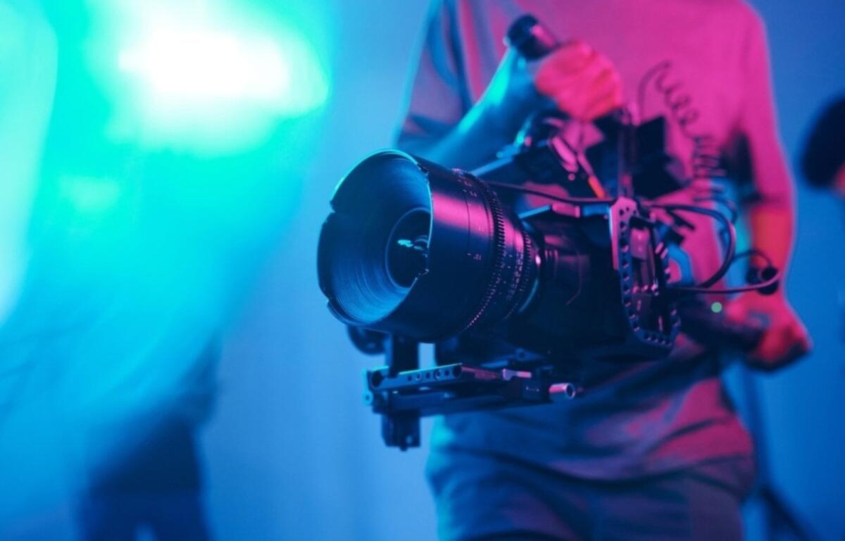 Cilmaker holding a professional video camera with a large lens on a stabilizing rig in a neon-lit studio.