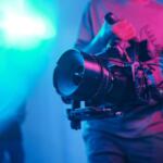 Cilmaker holding a professional video camera with a large lens on a stabilizing rig in a neon-lit studio.