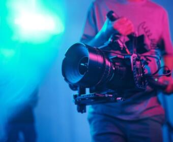Cilmaker holding a professional video camera with a large lens on a stabilizing rig in a neon-lit studio.