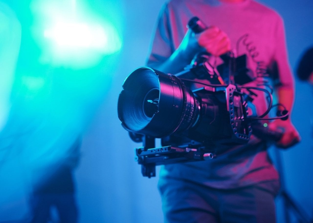 Cilmaker holding a professional video camera with a large lens on a stabilizing rig in a neon-lit studio.