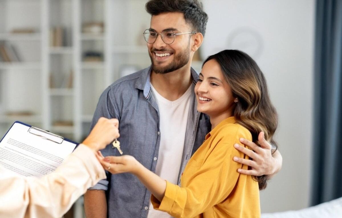 Couple smiles as they receive house keys from a person with a clipboard in a bright living room, celebrating a new home.
