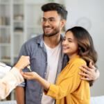 Couple smiles as they receive house keys from a person with a clipboard in a bright living room, celebrating a new home.