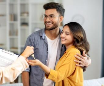 Couple smiles as they receive house keys from a person with a clipboard in a bright living room, celebrating a new home.