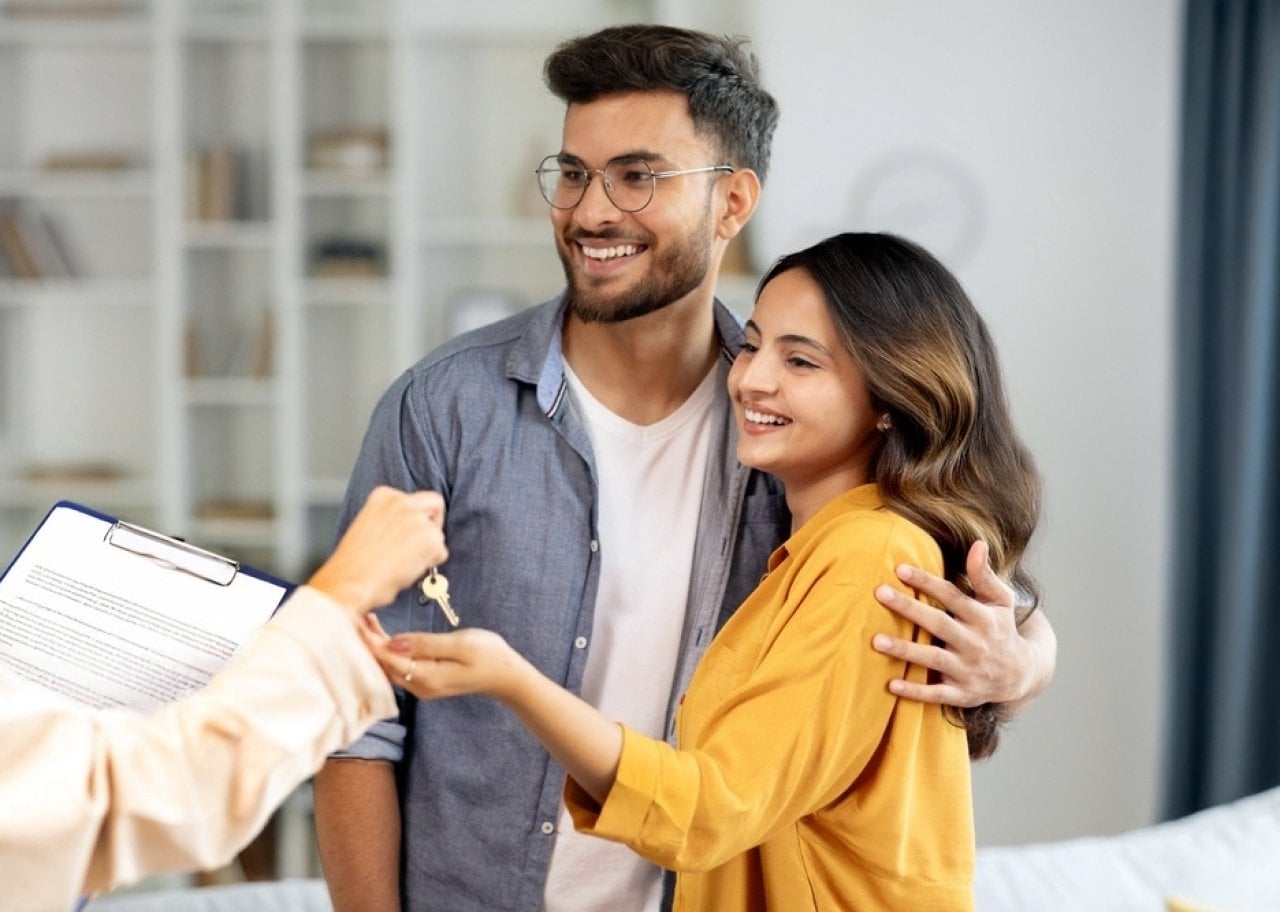 Couple smiles as they receive house keys from a person with a clipboard in a bright living room, celebrating a new home.