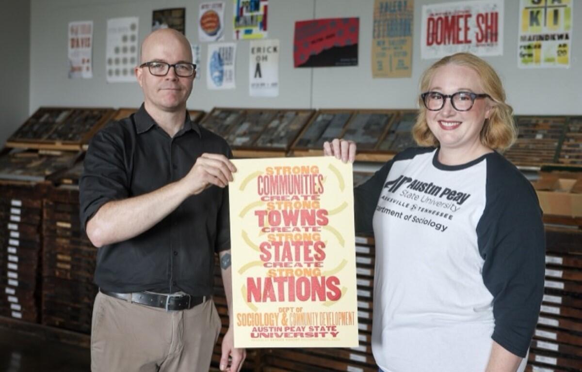 Two adults stand side by side and hold a poster promoting strong communities and nations at a university setting.