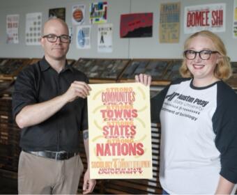 Two adults stand side by side and hold a poster promoting strong communities and nations at a university setting.