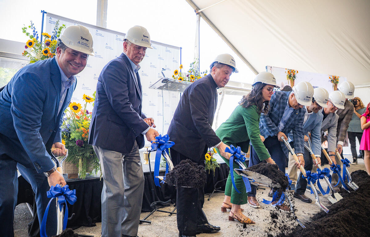 Group of people in hard hats and business attire shovel dirt at a groundbreaking ceremony under a tent, with blue ribbons on the shovels.