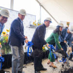 Group of people in hard hats and business attire shovel dirt at a groundbreaking ceremony under a tent, with blue ribbons on the shovels.