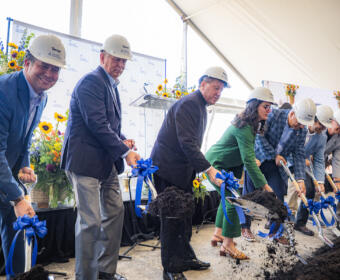 Group of people in hard hats and business attire shovel dirt at a groundbreaking ceremony under a tent, with blue ribbons on the shovels.