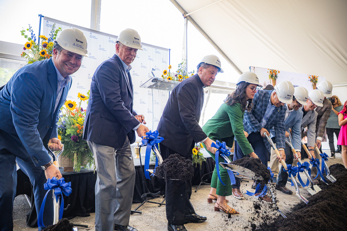 Group of people in hard hats and business attire shovel dirt at a groundbreaking ceremony under a tent, with blue ribbons on the shovels.