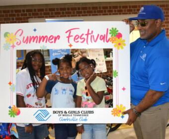 Three girls pose inside a colorful 'Summer Festival' frame with a man in a blue polo at a Boys & Girls Clubs event in Clarksville, Tennessee.