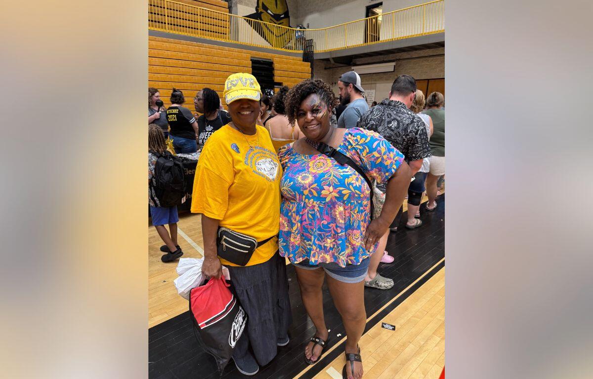 Two women smile for a photo in a gymnasium, one in a yellow shirt and cap, the other in a bright floral top with shorts and sandals, surrounded by people.