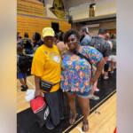 Two women smile for a photo in a gymnasium, one in a yellow shirt and cap, the other in a bright floral top with shorts and sandals, surrounded by people.