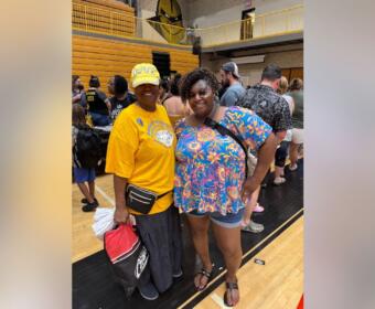 Two women smile for a photo in a gymnasium, one in a yellow shirt and cap, the other in a bright floral top with shorts and sandals, surrounded by people.