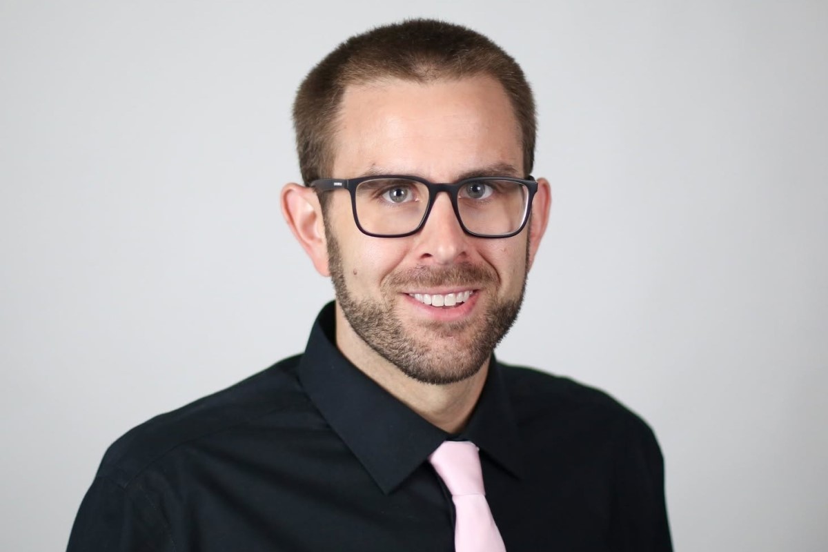 Head-and-shoulders portrait of a smiling man with short hair and glasses, wearing a black shirt and pink tie against a light gray background.
