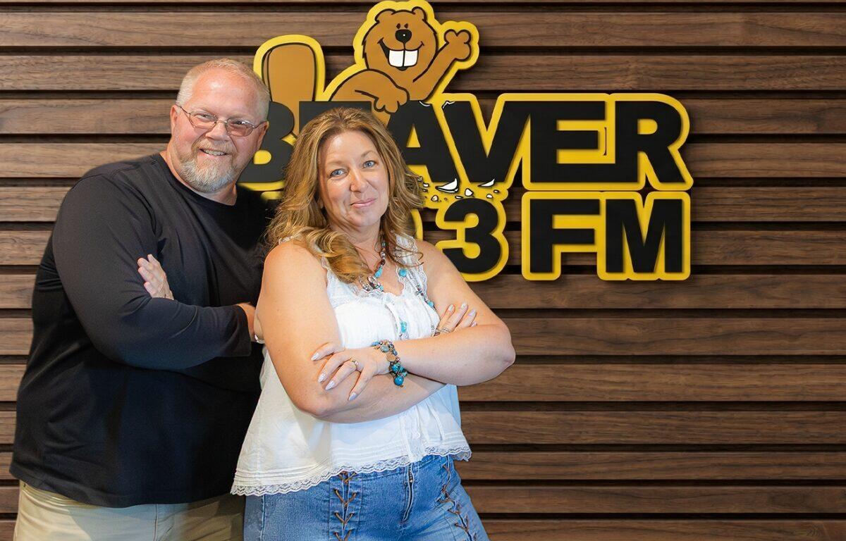 Two adults with arms crossed pose in front of a wood-paneled wall and a Beaver 3 FM sign with a cartoon beaver mascot above them.