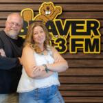 Two adults with arms crossed pose in front of a wood-paneled wall and a Beaver 3 FM sign with a cartoon beaver mascot above them.