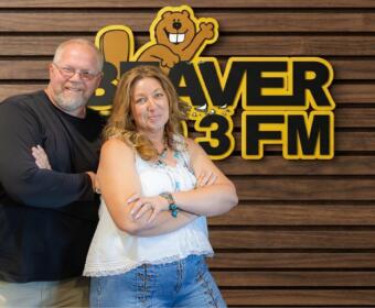 Two adults with arms crossed pose in front of a wood-paneled wall and a Beaver 3 FM sign with a cartoon beaver mascot above them.