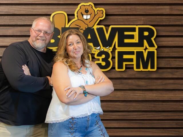 Two adults with arms crossed pose in front of a wood-paneled wall and a Beaver 3 FM sign with a cartoon beaver mascot above them.