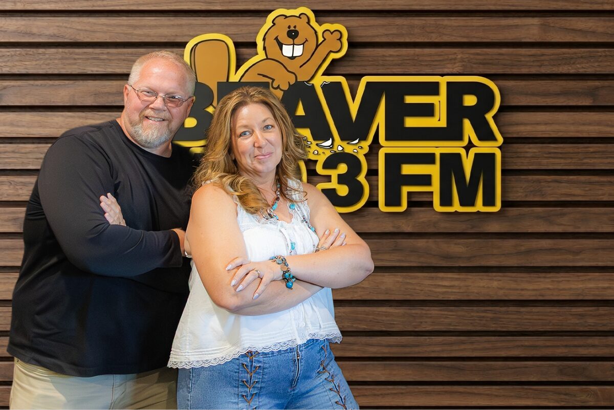 Two adults with arms crossed pose in front of a wood-paneled wall and a Beaver 3 FM sign with a cartoon beaver mascot above them.