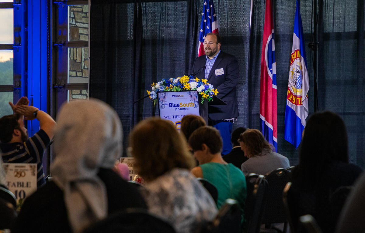Speaker at a podium with blue floral arrangement, giving a talk at a banquet with flags behind him.