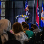Speaker at a podium with blue floral arrangement, giving a talk at a banquet with flags behind him.