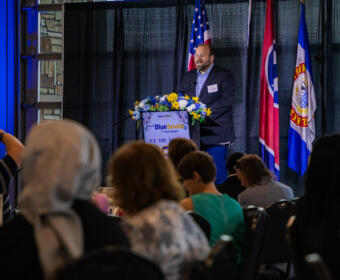 Speaker at a podium with blue floral arrangement, giving a talk at a banquet with flags behind him.