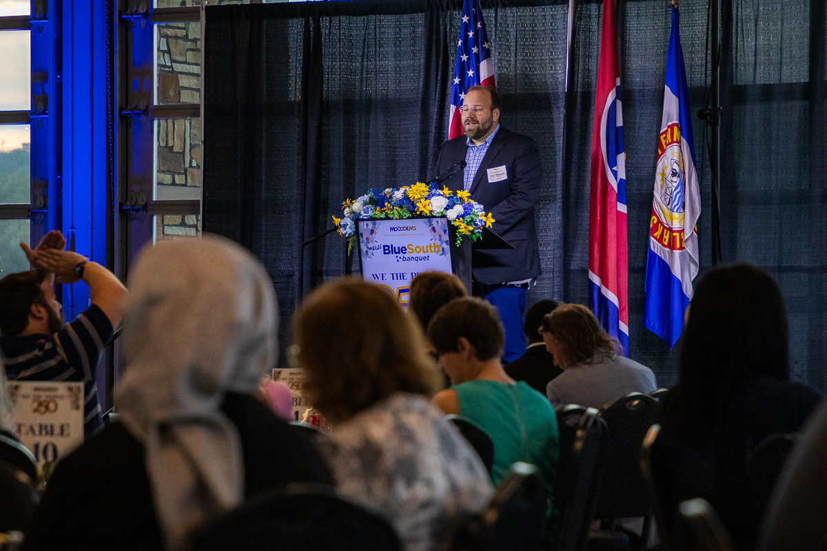 Speaker at a podium with blue floral arrangement, giving a talk at a banquet with flags behind him.