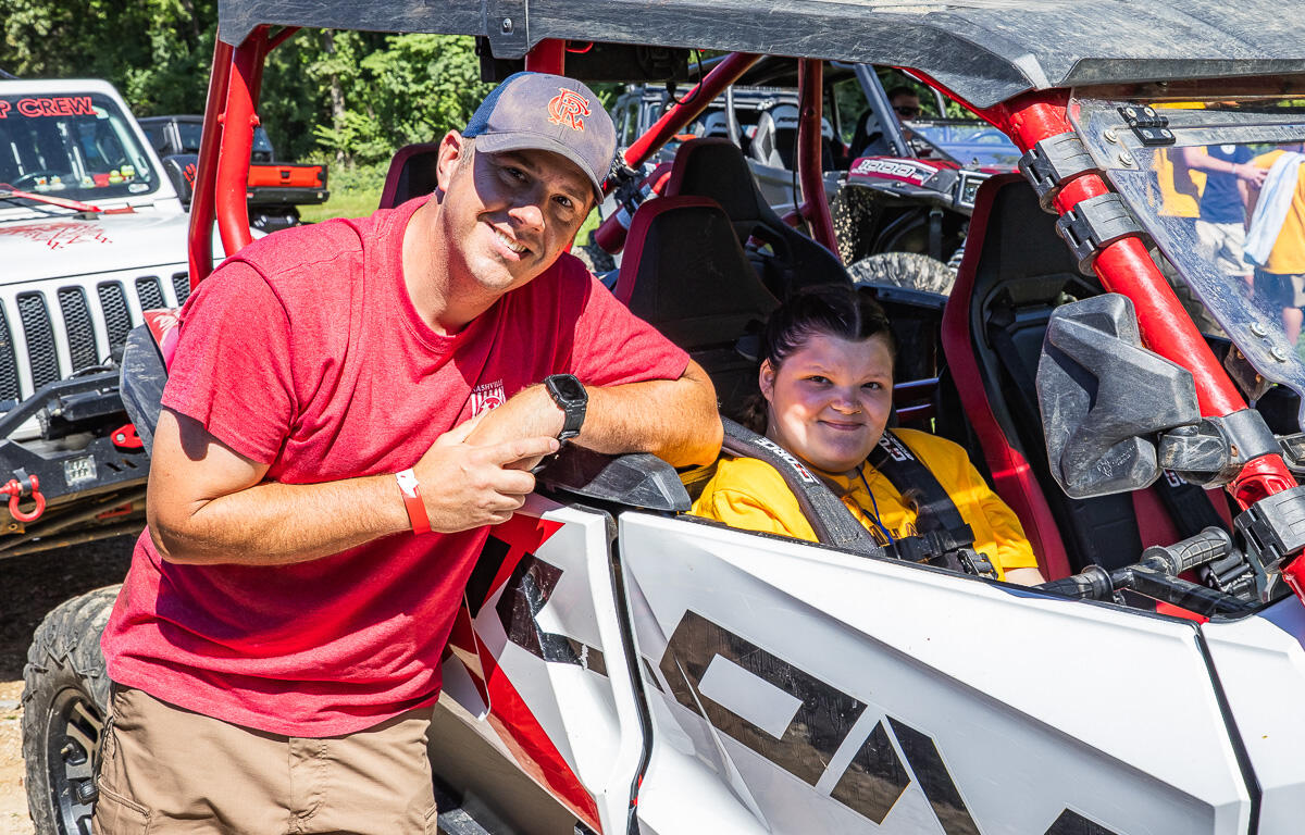 Man in red shirt and boy/girl in yellow sit in a white and red off-road dune buggy, posing for the camera beside the vehicle.