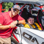 Man in red shirt and boy/girl in yellow sit in a white and red off-road dune buggy, posing for the camera beside the vehicle.
