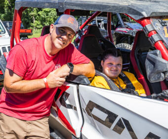 Man in red shirt and boy/girl in yellow sit in a white and red off-road dune buggy, posing for the camera beside the vehicle.