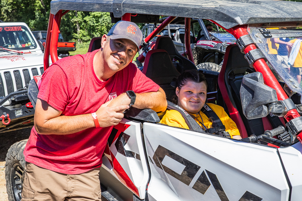 Man in red shirt and boy/girl in yellow sit in a white and red off-road dune buggy, posing for the camera beside the vehicle.