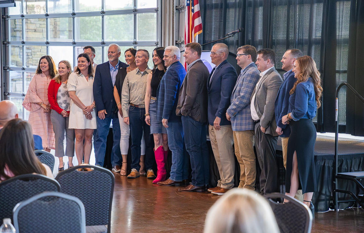 Group of adults posing for a photo on a stage in a conference hall, with an American flag in the background and audience chairs in the foreground.