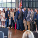 Group of adults posing for a photo on a stage in a conference hall, with an American flag in the background and audience chairs in the foreground.