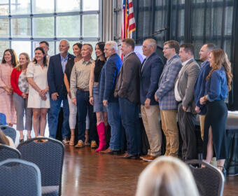 Group of adults posing for a photo on a stage in a conference hall, with an American flag in the background and audience chairs in the foreground.