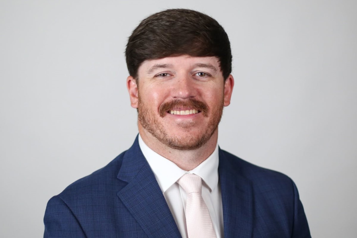 Head-and-shoulders portrait of a smiling man in a blue suit and light pink tie against a light gray background.