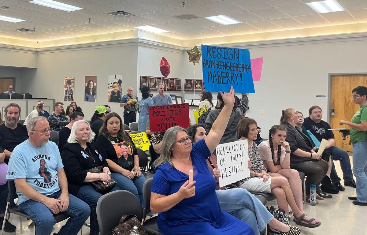 Audience seated in a meeting room, several people holding protest signs, including a blue sign that says 'RESIGN' and a white sign calling for resignation.