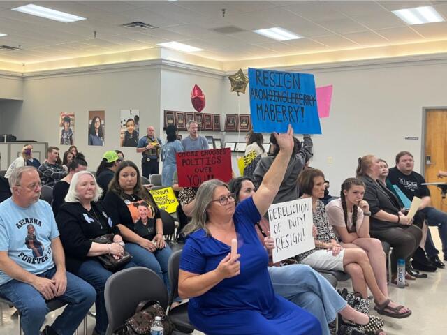 Audience seated in a meeting room, several people holding protest signs, including a blue sign that says 'RESIGN' and a white sign calling for resignation.