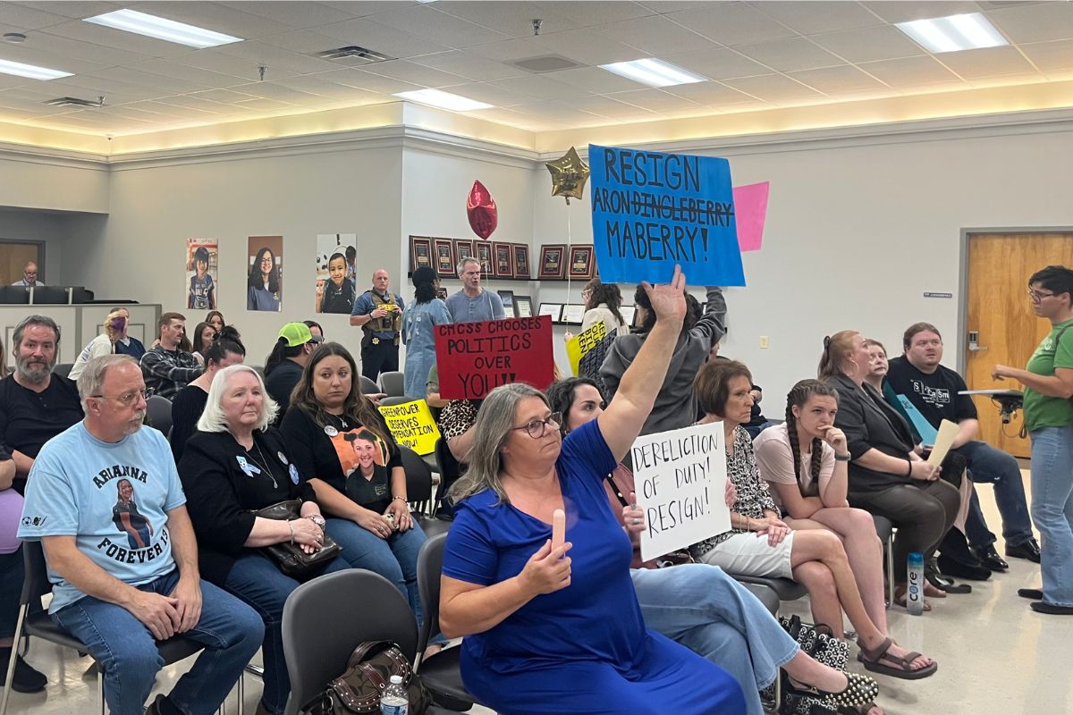 Audience seated in a meeting room, several people holding protest signs, including a blue sign that says 'RESIGN' and a white sign calling for resignation.
