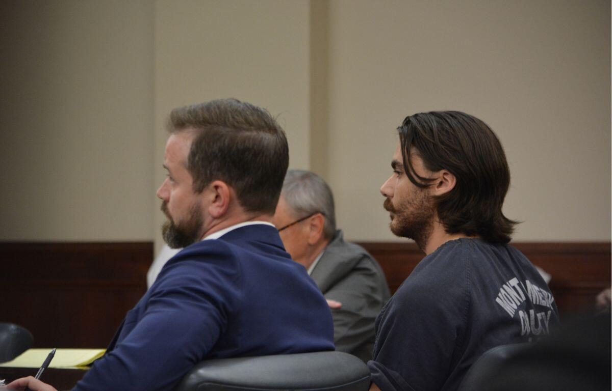 Two men sit side by side in a courtroom, one in a blue suit and the other with long hair in a dark shirt, facing forward.