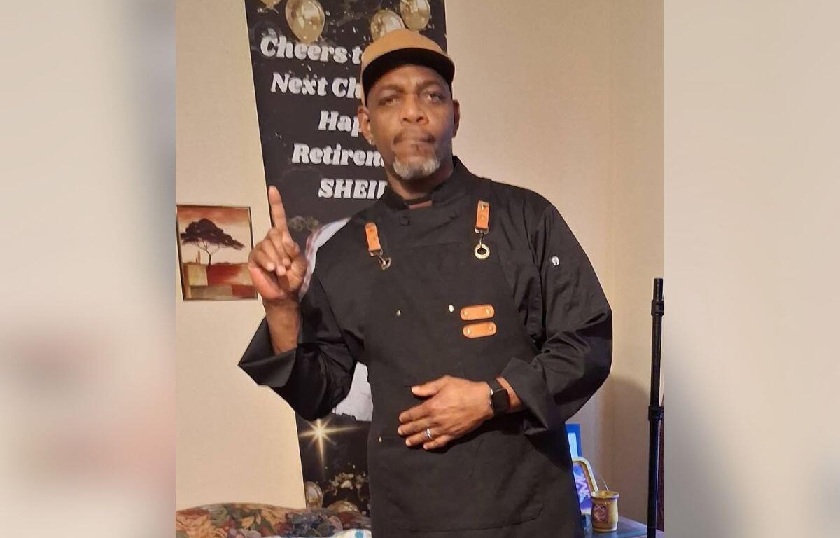 Man in a black chef jacket and tan cap stands indoors, pointing upward with his right finger as if making a point.