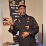 Man in a black chef jacket and tan cap stands indoors, pointing upward with his right finger as if making a point.