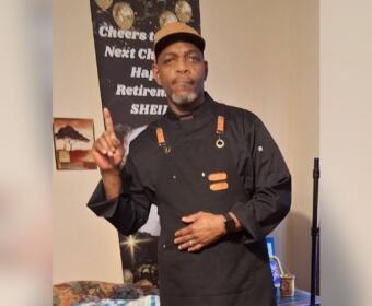 Man in a black chef jacket and tan cap stands indoors, pointing upward with his right finger as if making a point.