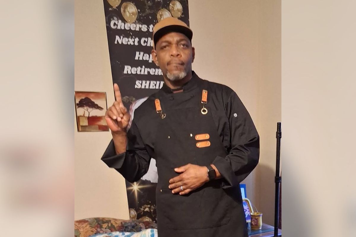 Man in a black chef jacket and tan cap stands indoors, pointing upward with his right finger as if making a point.