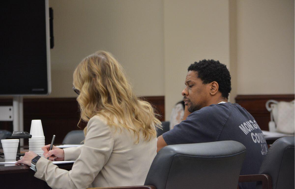 Two people sit at a conference table in a formal room; a blonde woman in a beige jacket writes in a notebook while a man in a dark T-shirt sits beside her.