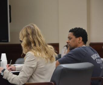 Two people sit at a conference table in a formal room; a blonde woman in a beige jacket writes in a notebook while a man in a dark T-shirt sits beside her.